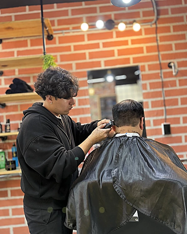 Un barbero de sudadera negra pasa la máquina por la nuca de un cliente de espaldas, frente al muro de ladrillo