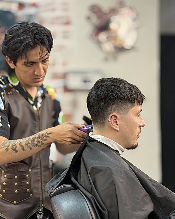 Barbero con chaleco de cuero y brazo tatuado marca el taper con la máquina detrás de la oreja de un cliente