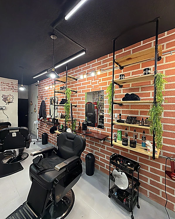 El salón a lo largo: sillones de barbero negros, el muro de ladrillo con repisas de madera, plantas y espejos, y el piso gris pulido