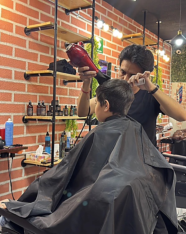 Un barbero seca y peina con secadora el corte de un niño de espaldas, frente a las repisas con productos