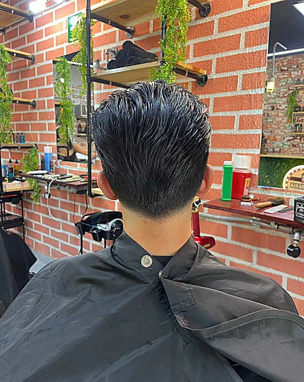 Corte visto desde la nuca: textura peinada hacia atrás con brillo y taper limpio en la nuca, con el muro de ladrillo y las repisas al fondo